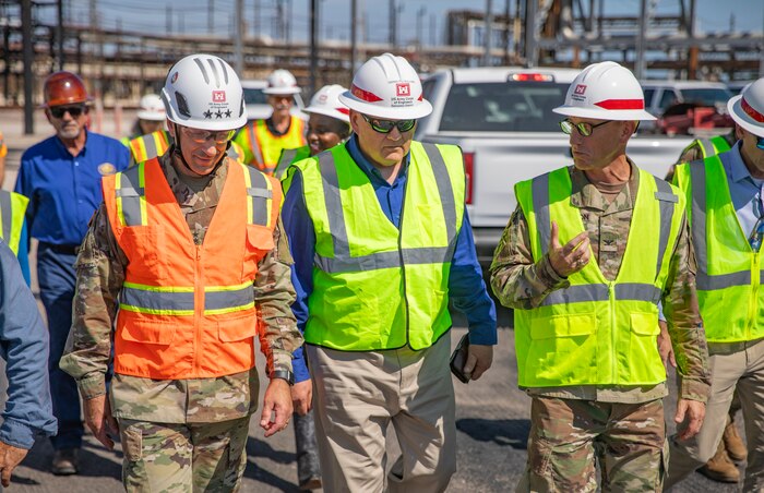 people in hard hats and safety vests are walking through an oil refinery, talking.
