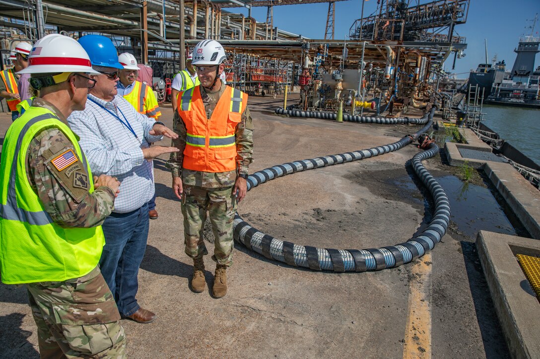 people in hard hats and safety vests are standing in an oil refinery near water, talking.