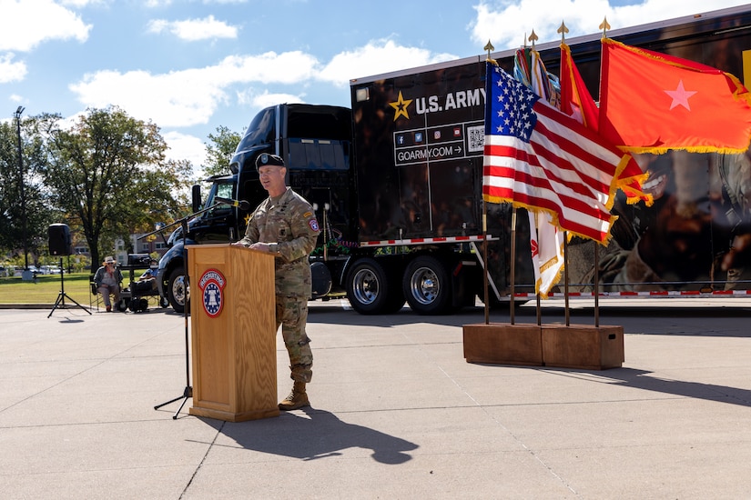 A man in US Army uniform stands behind a podium outside.