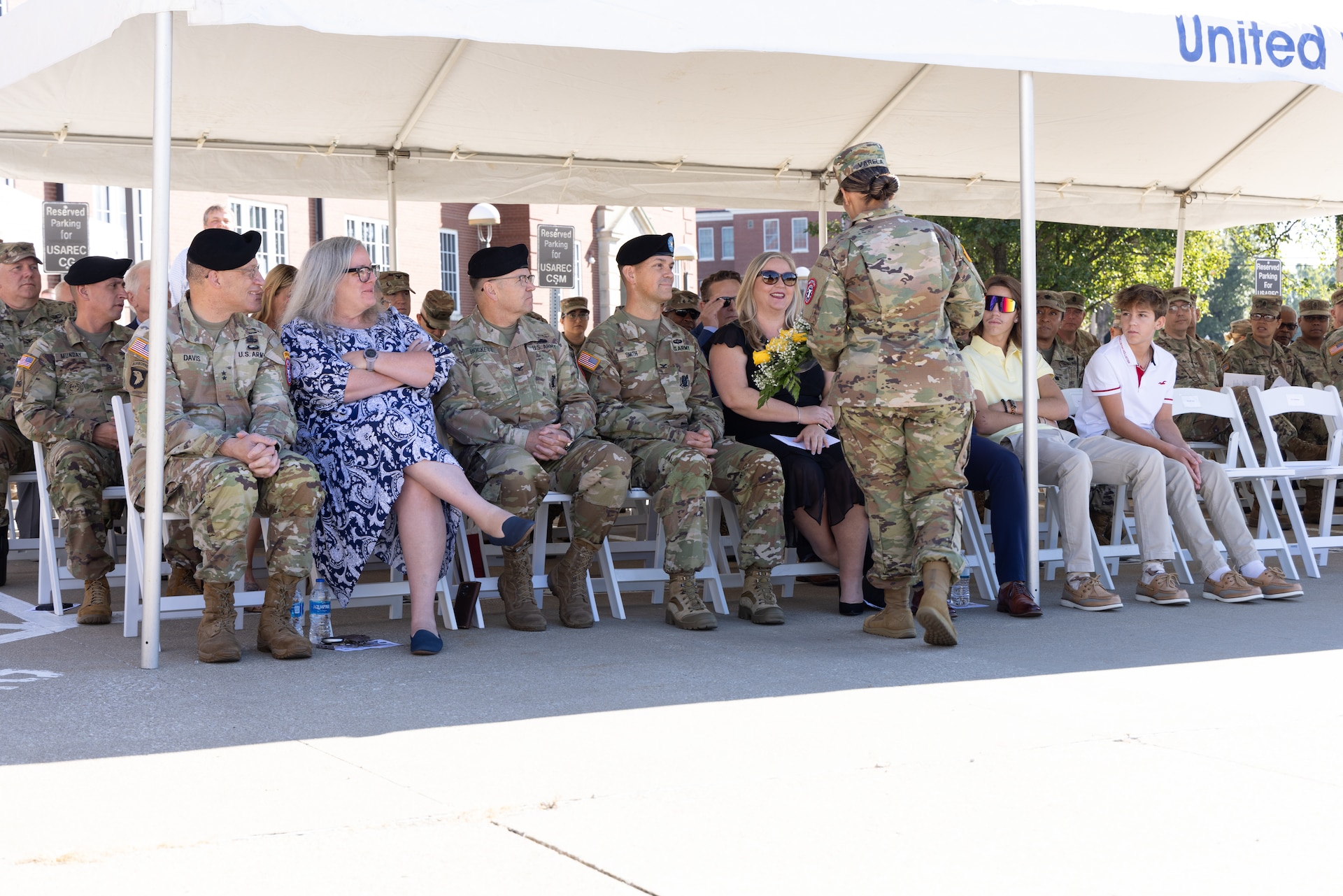 Group of soldiers and civilians sit under a tent outside.