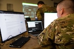 A soldier in uniform is seated at a computer workstation.