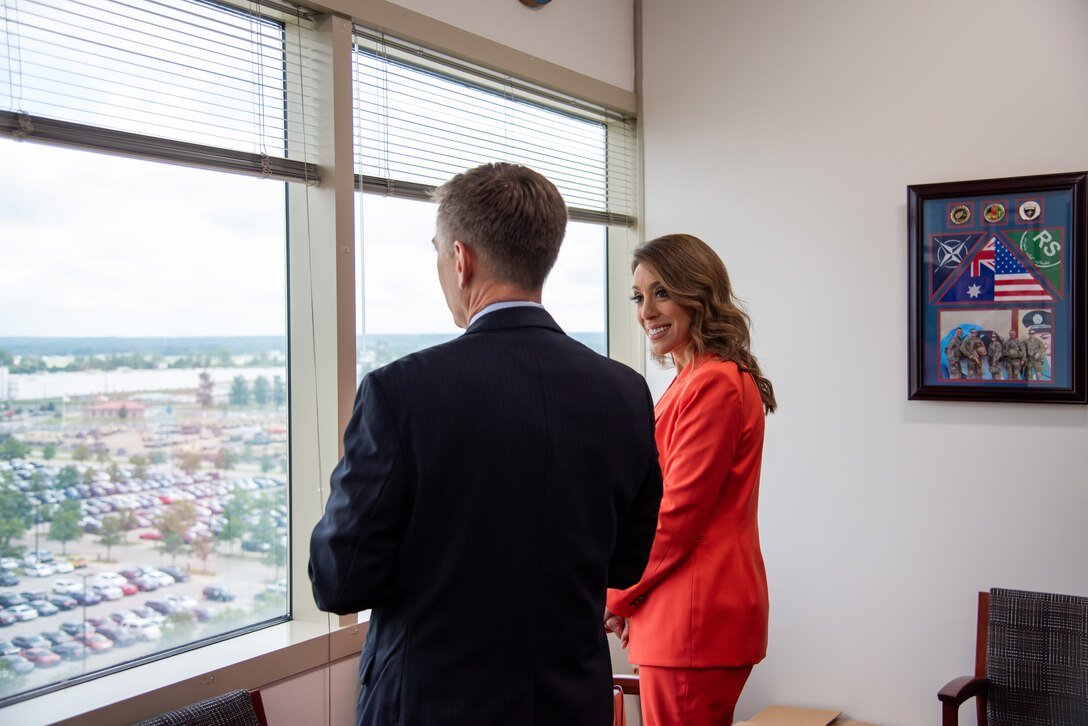 Man in suit and woman in orange suit stand looking out a window.