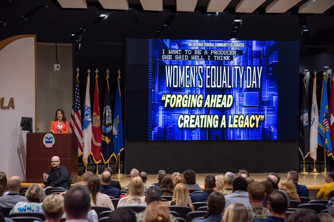 Woman on stage stands behind podium, 'Women's Equality Day' on screen behind.