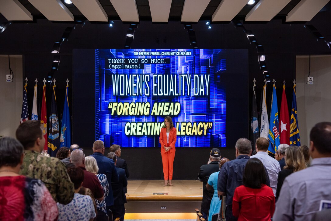 Women wearing orange suit stands on stage with 'Women's Equality Day' on screen behind.