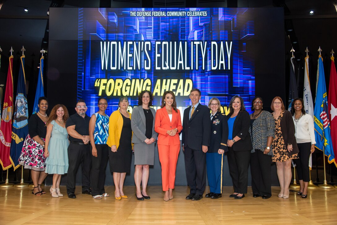 Thirteen people pose standing on stage with 'Women's Equality Day' on screen behind.