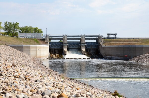 rocks on the side of water, bridge and dam gates above, blue sky, green trees