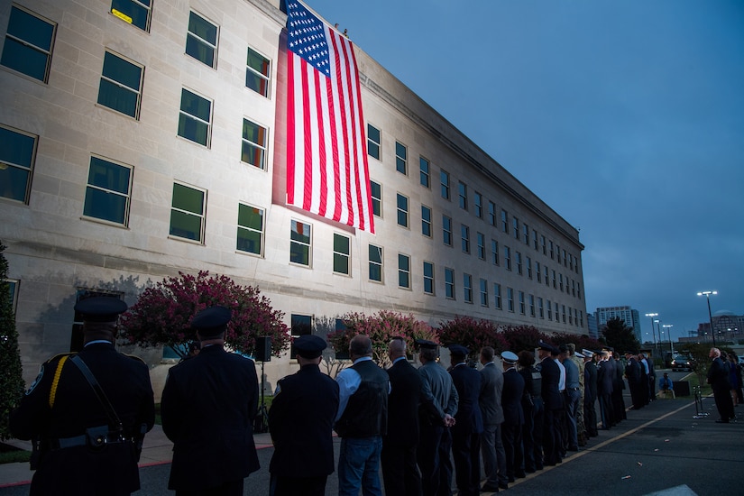 People line up at dawn to look at a flag in a spotlight that’s unfurled onto a large building.