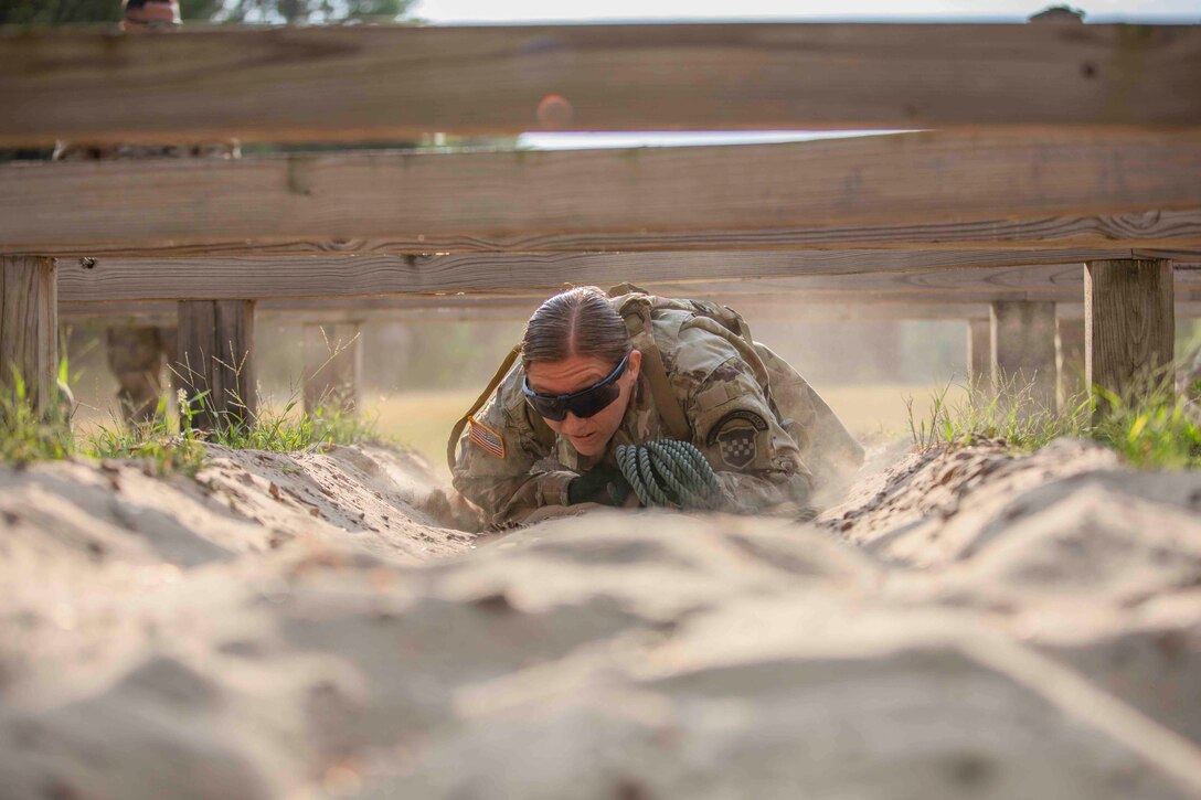 A soldier low-crawls on sand underneath an obstacle.