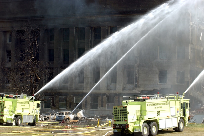 Two fire rescue vehicles and a stand-alone cannon spray fire-suppressant material onto a large, damaged building.