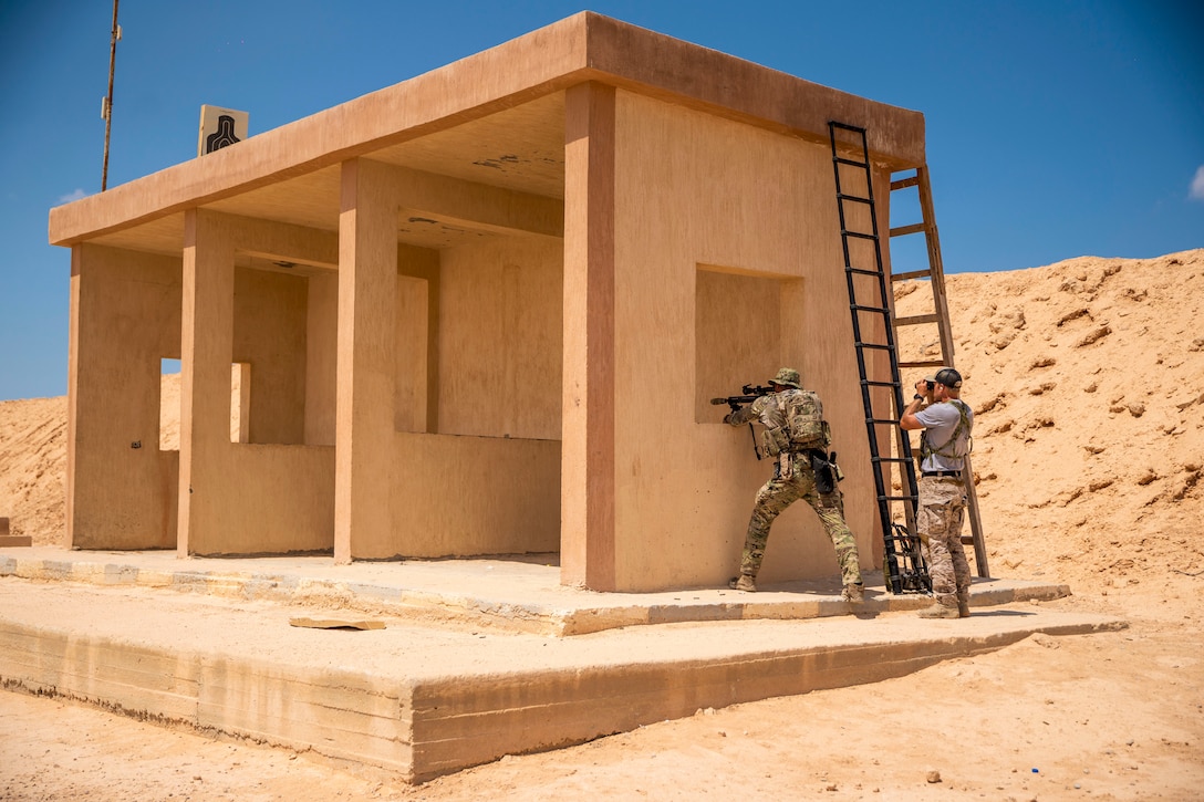 A sailor aims a weapon through the first of four squares in a wooden structure in a desert-like area as a fellow service member watches through binoculars.