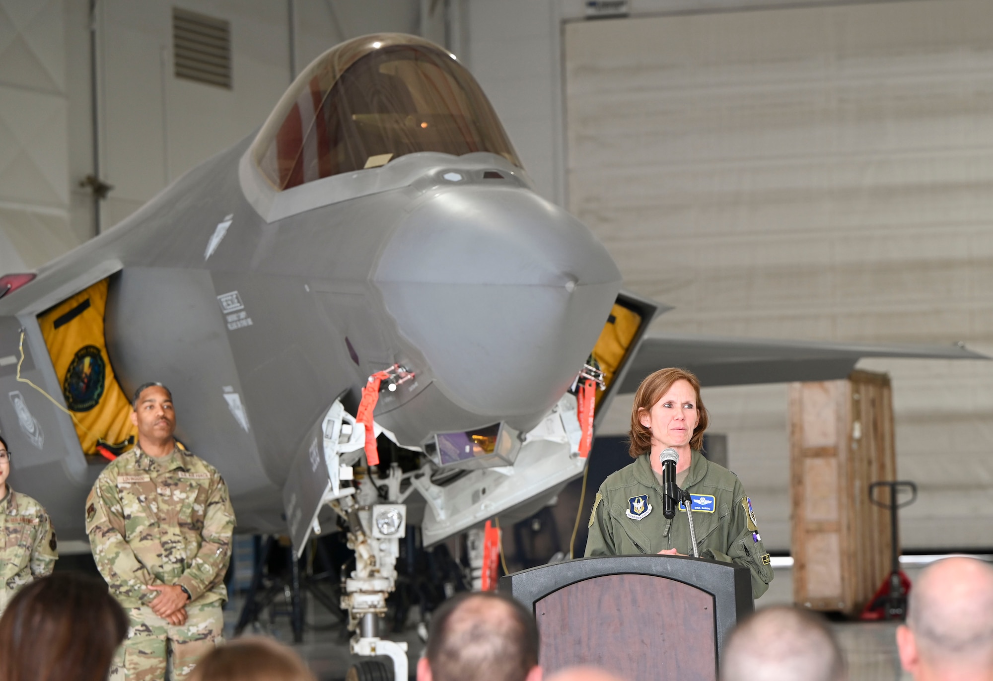 Brig. Gen. Regina Sabric speaks while standing at a podium. An F-35 aircraft is staged behind her.