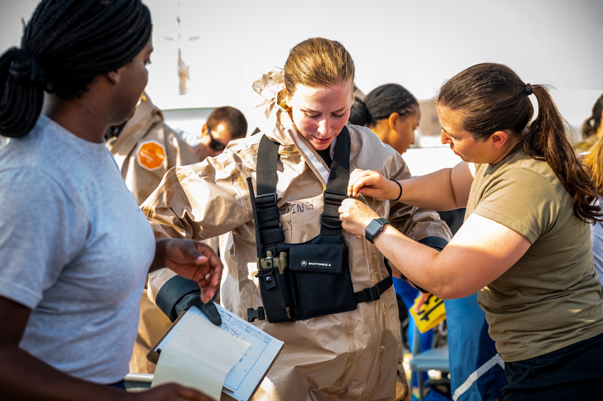 Military member in chemical gear receives help taking off her equipment.