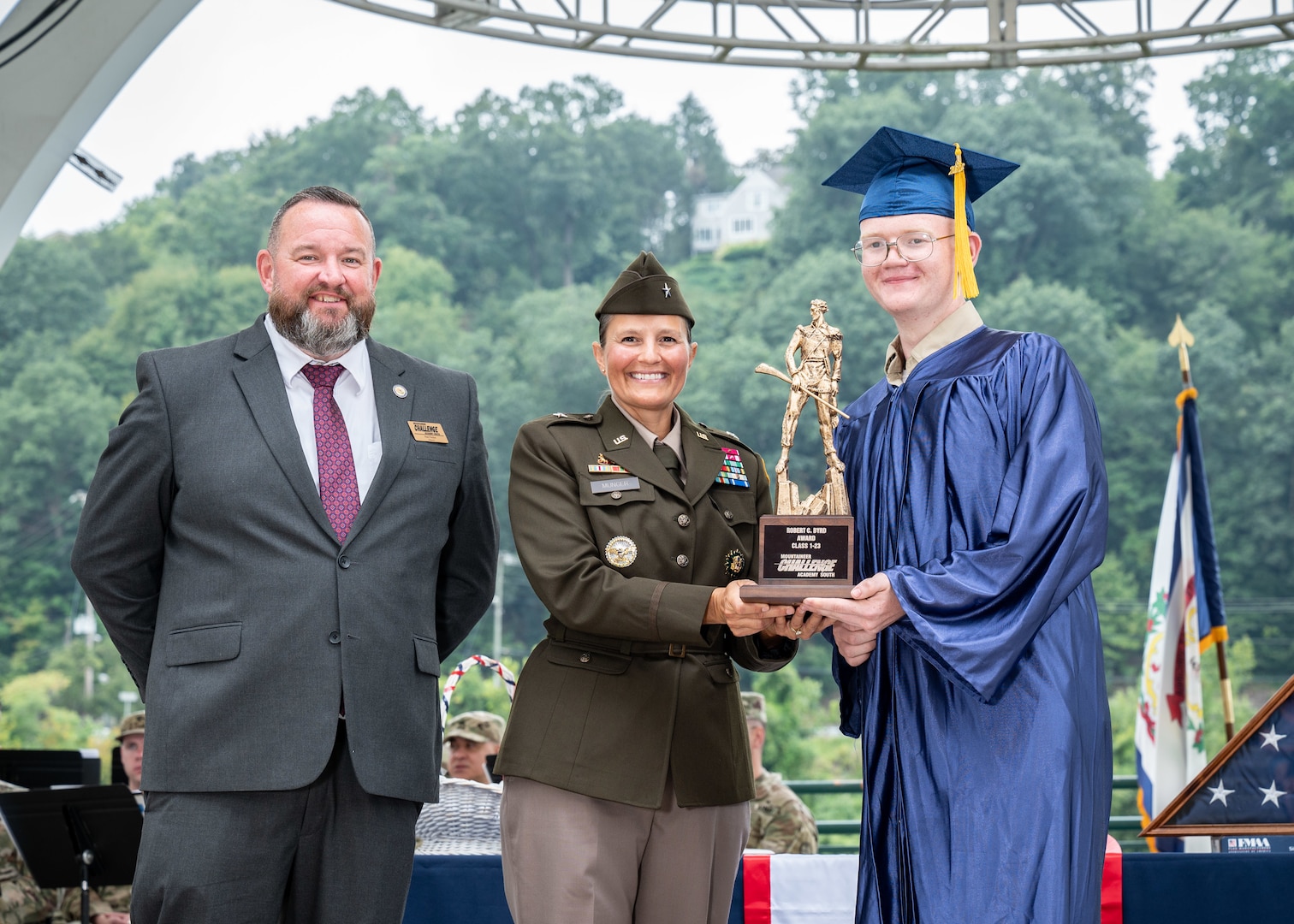 a close-up of three people looking at the camera in a portrait. a white teen (right) is wearing a blue graduation robe and hat. He's holding half of the base if a gold statue of a mountaineer man (about 1 foot tall) that's on a wood base. A woman (center) in U.S. Army green dress uniform is holding the other half. She has dark brown hair in a bun. A man with brown hair and beard is in a grey suit, standing on the left. All three are smiling.