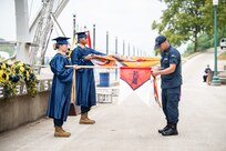 Profile shot of cadre (on right) switching guidon ribbons from company guidon to MCA-South flag guidon. A cadet in royal blue graduation robe and motor board hat is holding each flag horizontally to the ground. They are outside in an Ampitheater on a river.