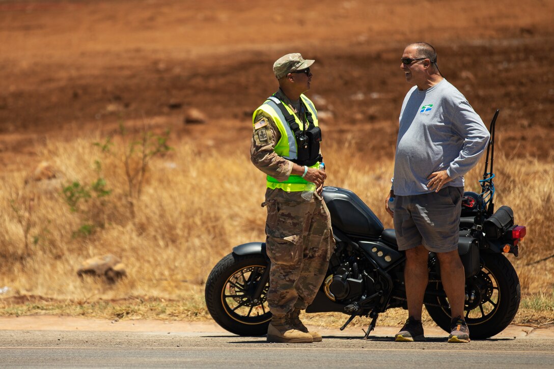 A soldier speaks to a resident next to a motorcycle on the side of the road.