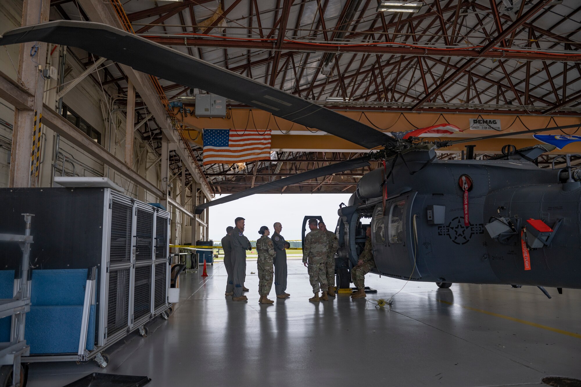 Airmen stand inside a hanger next to a large gray helicopter