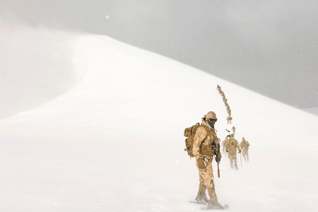 Marines wearing winter gear climb a snowy mountain in formation.