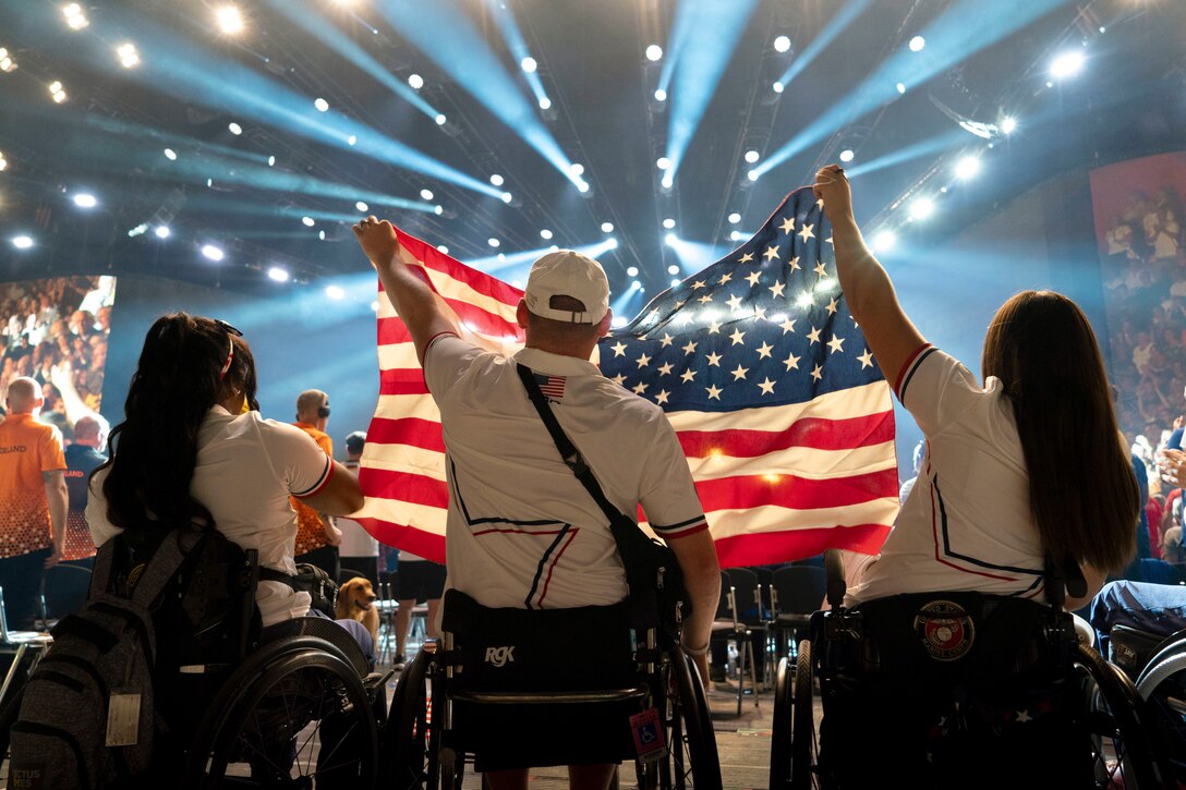 Three people in wheelchairs hold an American flag during a ceremony under spotlights.