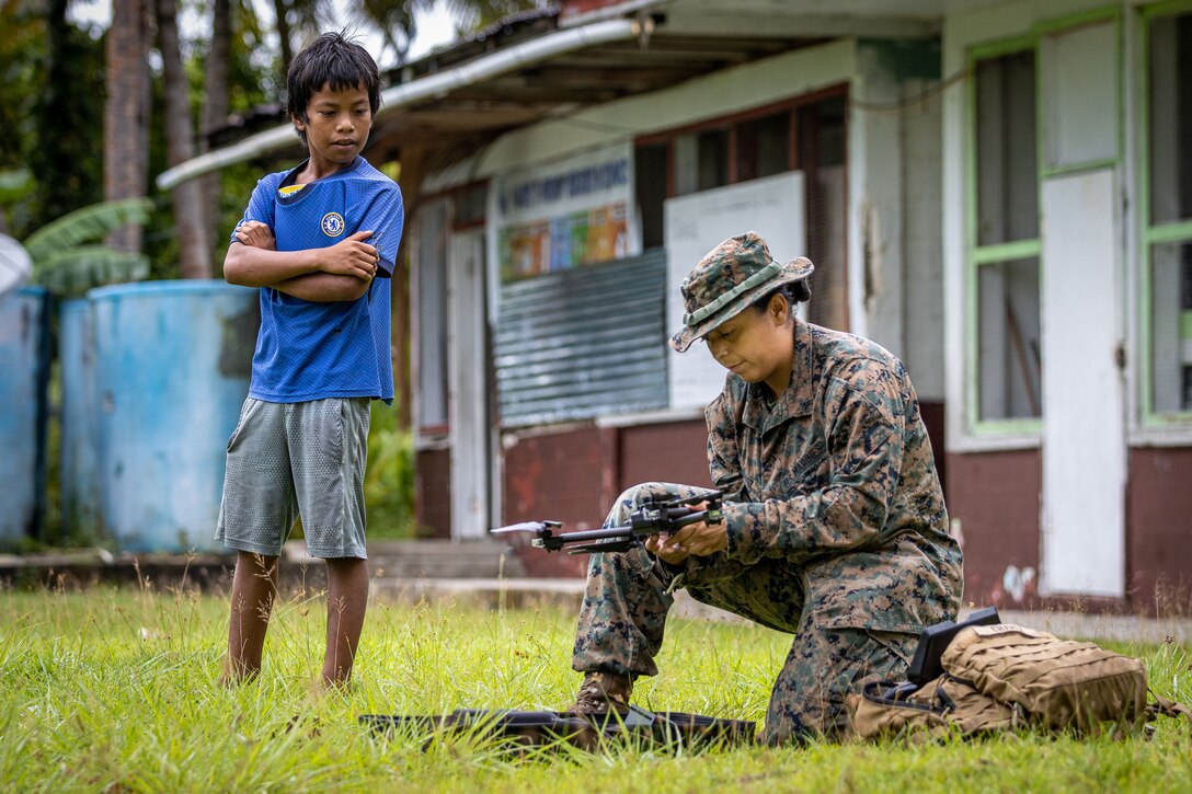 A Marine kneels on the grass while handling a drone as a child stands and watches.