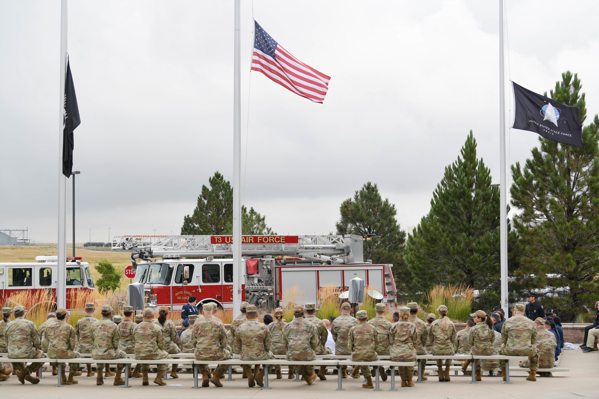Military members sit on benches and watch someone speak at a podium.