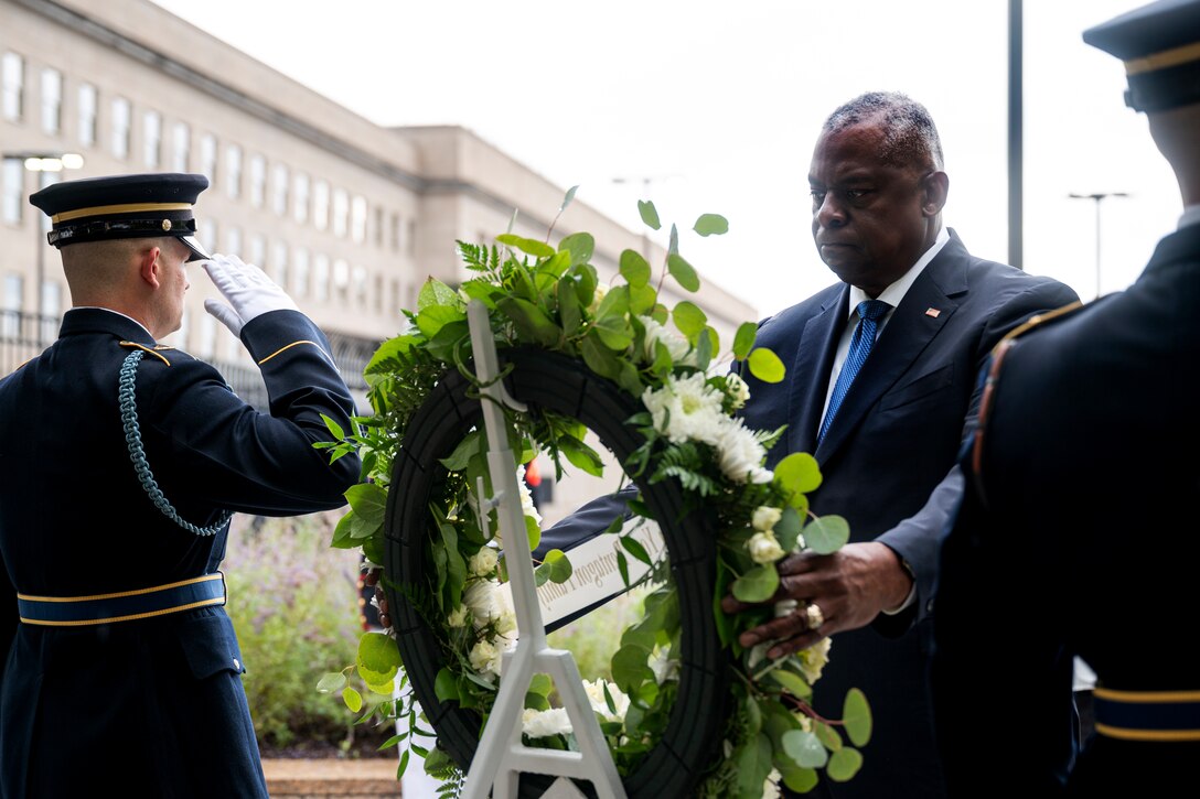 Secretary of Defense Lloyd J. Austin III places a wreath on a stand outside the Pentagon as a service member salutes.