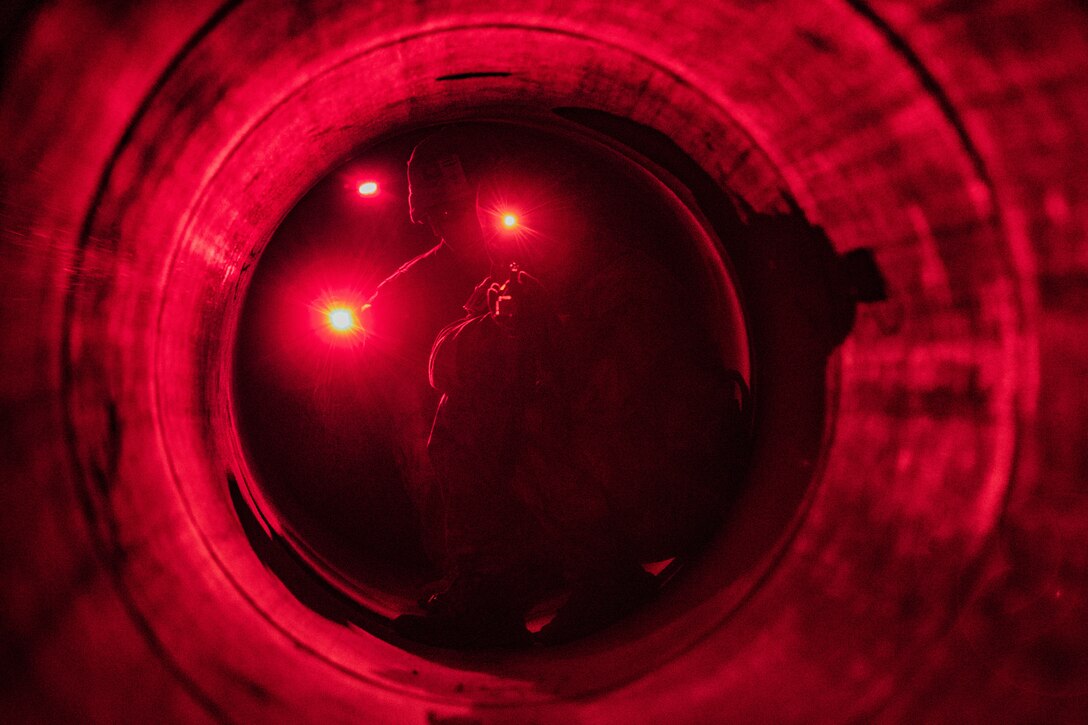 A soldier aims his gun down a tunnel lit by red lights.