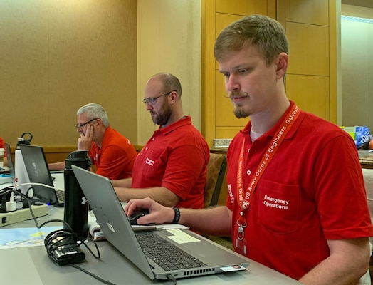 Three men in red shirts sit at a table working on laptop computers