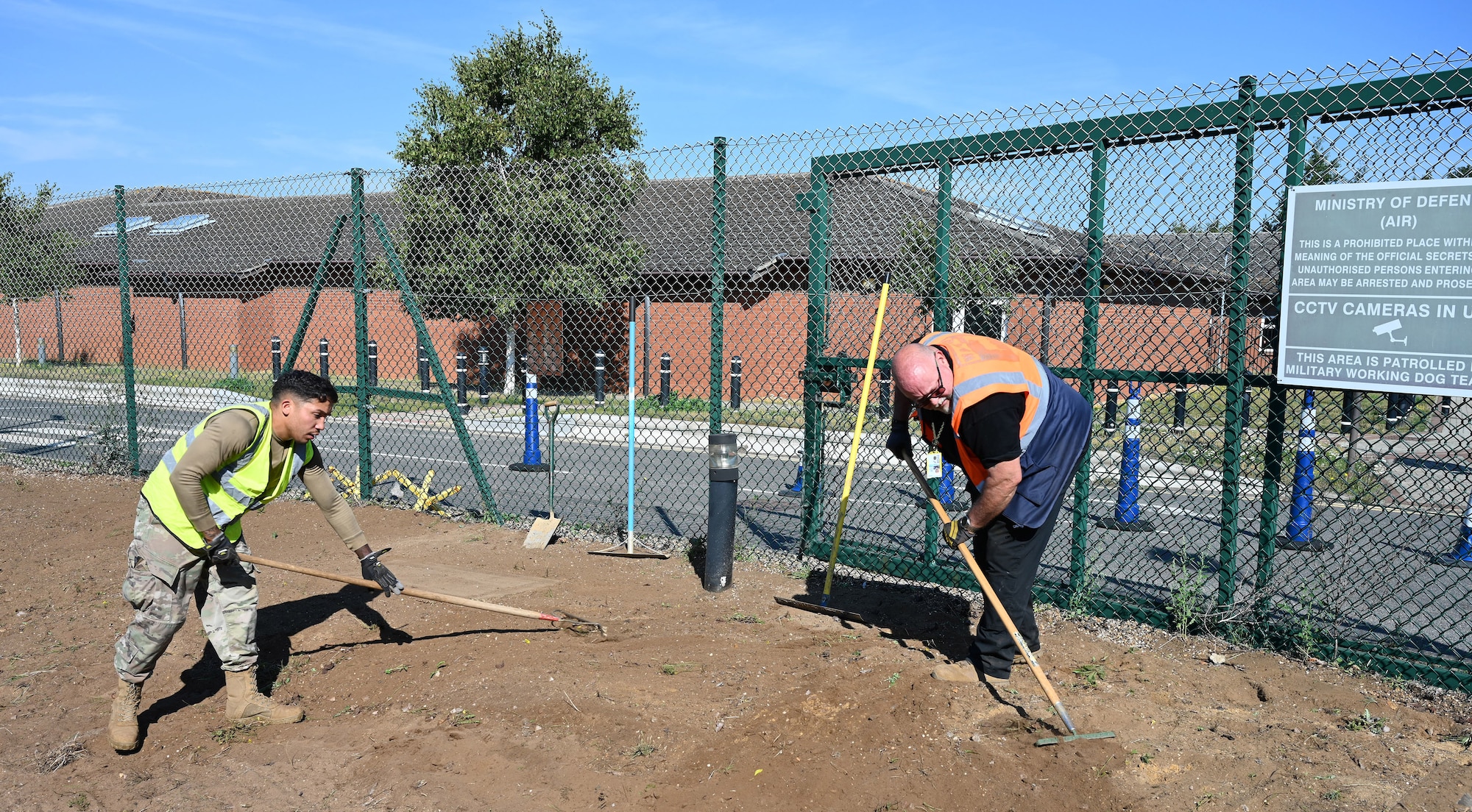 U.S. Air Force Staff Sgt. Isaiah Torres, left, 100th Civil Engineer Squadron Heavy Equipment and Pavements craftsman, and Paul Waites, 100th CES Heavy Equipment and Pavements foreman, rake soil to remove remaining weeds and stones after removing paving slabs next to the new pedestrian entry control point at the child development center at Royal Air Force Mildenhall, England, Sept. 7, 2023. As part of a base initiative to make the entrance area safer and more secure for CDC customers within the tri-base area, the heavy equipment and pavements Airmen, otherwise known as the “Dirt Boyz,” put up new fencing, replaced curbs and pathways, and added a concrete mini hardstand for an additional 100th Security Forces Squadron entry control point to provide easier and safer access for those walking across the main road from base to the CDC gate. (U.S. Air Force photo by Karen Abeyasekere)