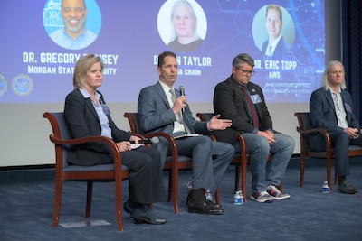 One woman and three men, all with microphones, sit in a row on a stage.