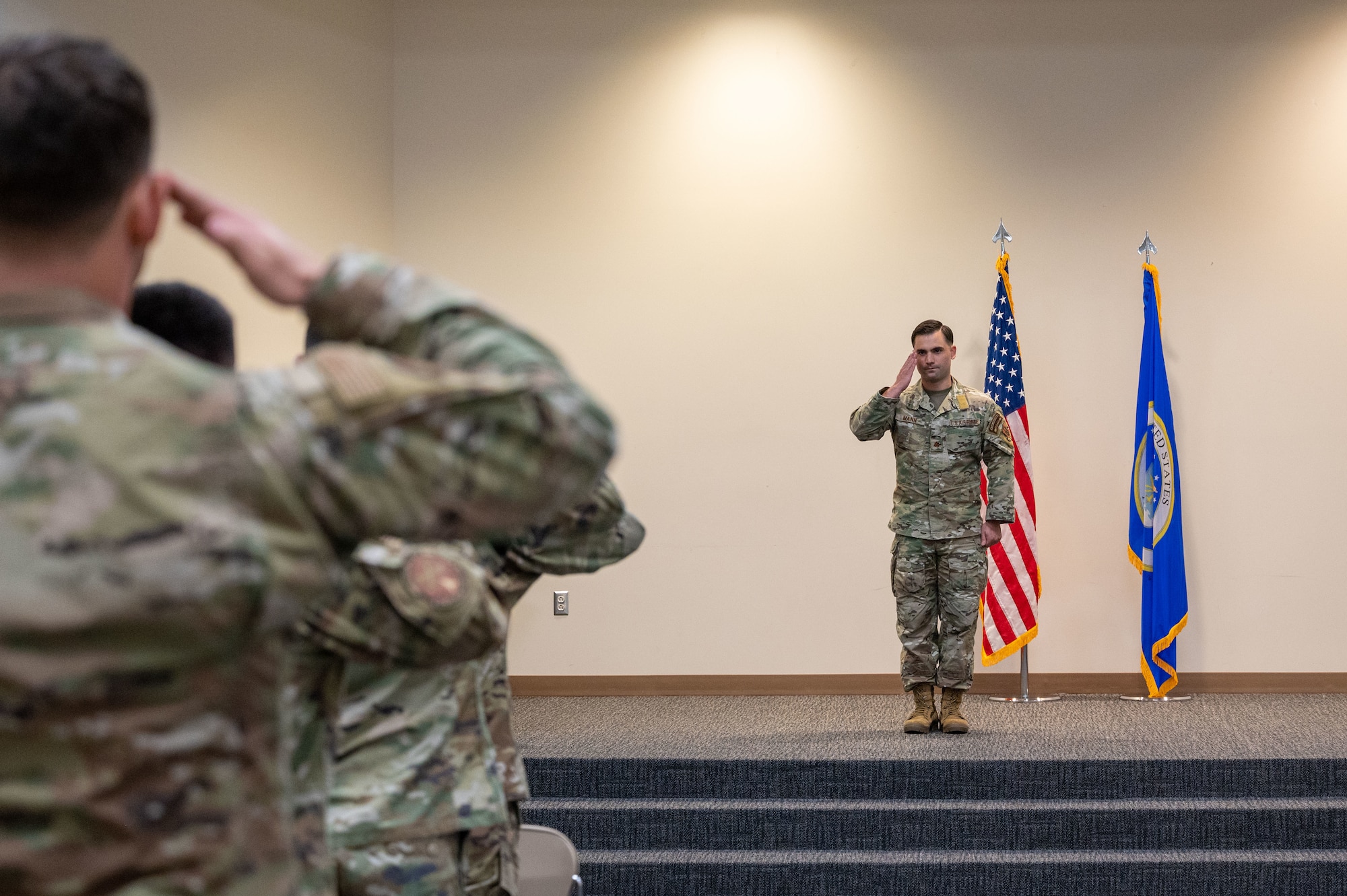 In the foreground, a group of uniformed members salute Mann who is standing on stage also saluting.