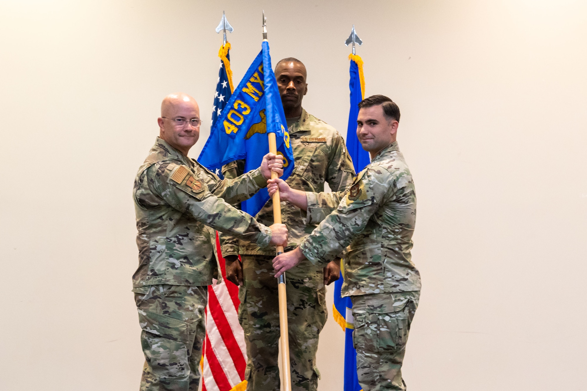 Three uniformed members stand flanked by the Air Force and American Flags. Magee on the left hands a guidon flag to Mann on the right.