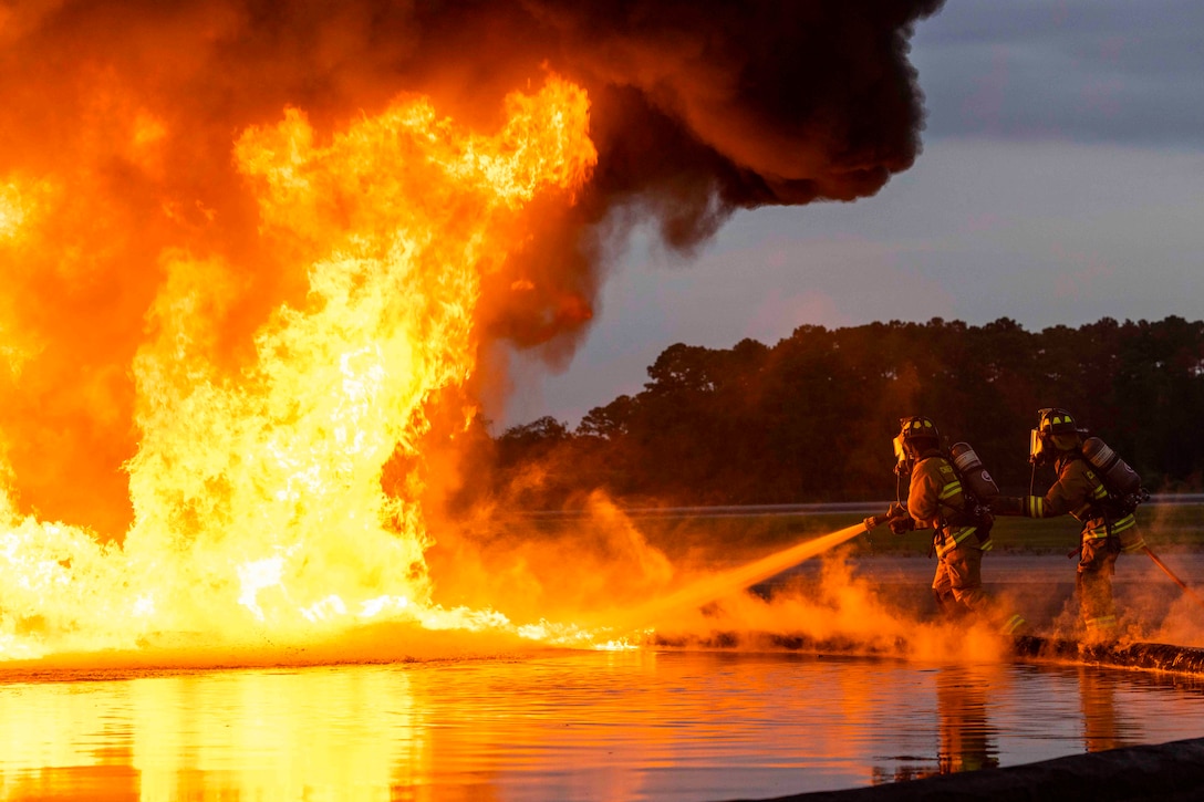 Two Marines wearing fire protection equipment use a hose to spray water onto a fire.