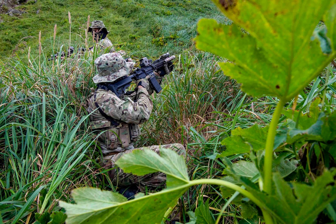 Two sailors move through vegetation with their weapons drawn.