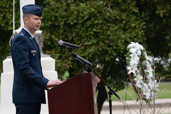 A Air Force member gives speech in uniform next to wreath with white flowers.