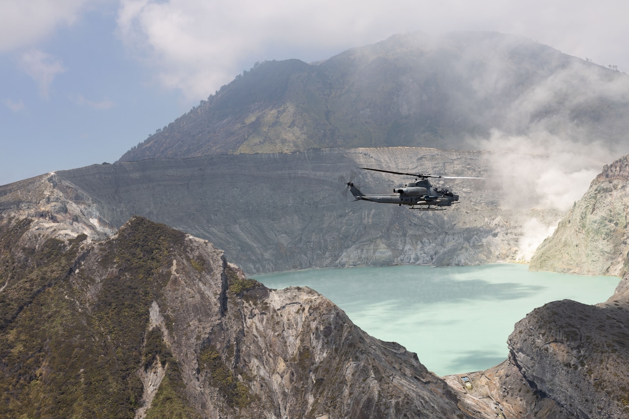 A military helicopter flies above mountainous terrain.