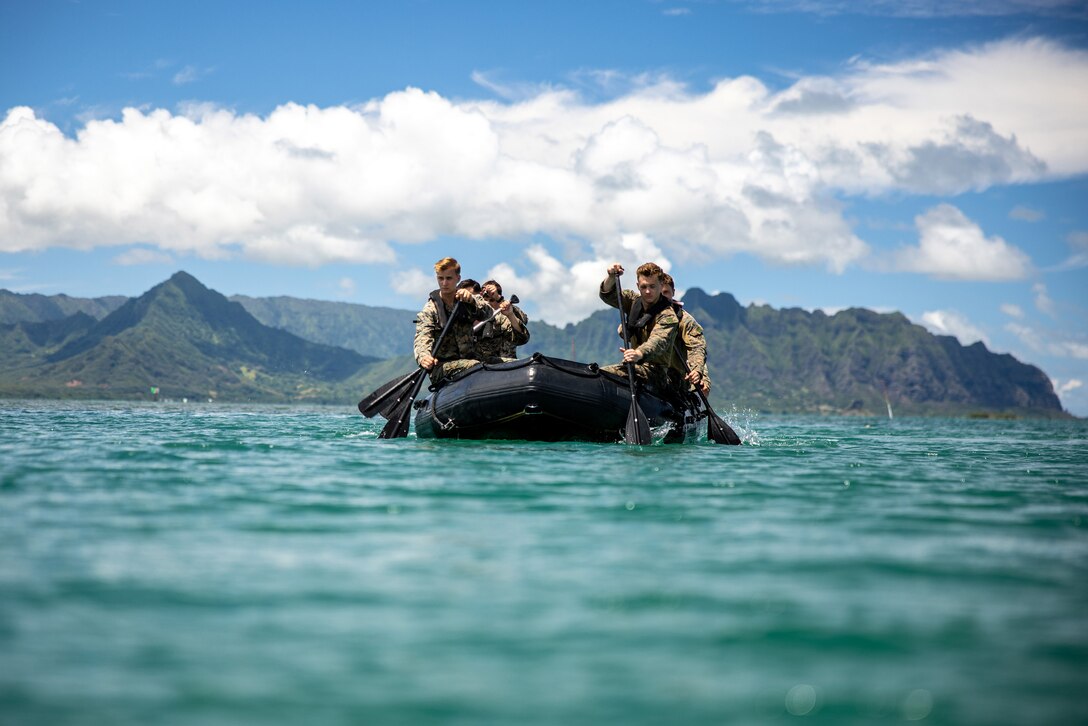 Marines ride in a rubber raft in open water during daylight with mountains behind them.
