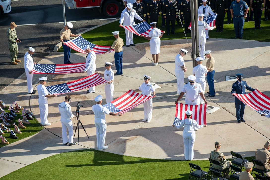 Sailors stand beneath a flagpole while holding flags during a ceremony.
