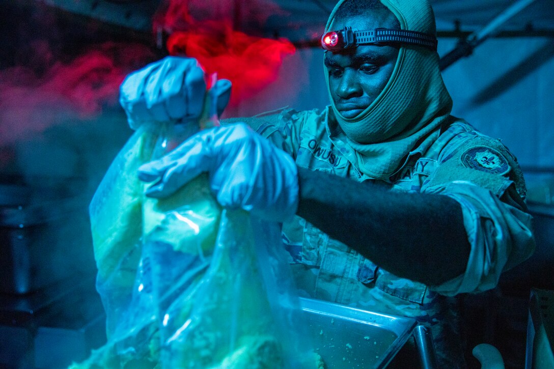 A soldier wearing a headlamp pours eggs from a plastic bag into a metal container illuminated by a red light.