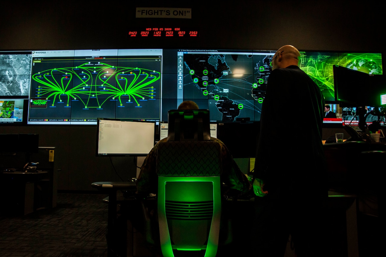 A seated person operates a computer. Another person stands nearby. Large video screens loom in the background.