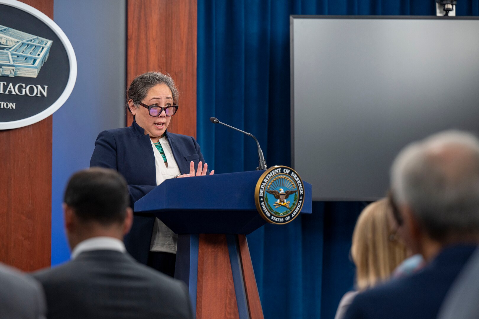 A civilian stands behind a lectern. Others are seated in front of her.