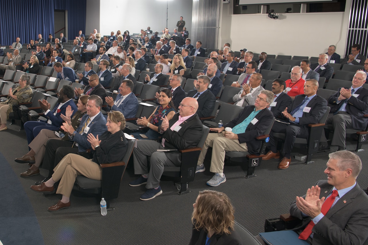 A large audience, wearing business attire, sits in an auditorium.