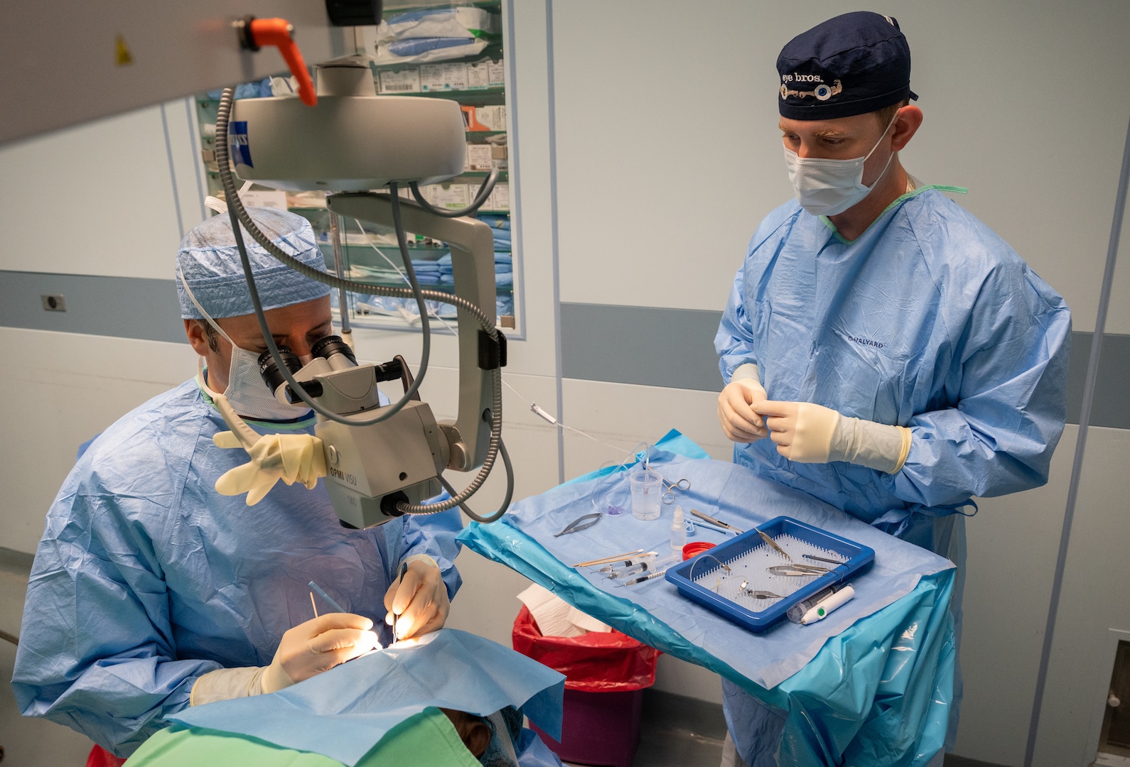 U.S. Air Force Col. (Dr.) Richard Townley, ophthalmologist at Joint Base Elmendorf- Richardson, Alaska, performs manual small incision cataract surgery on a patient while U.S. Air Force Maj. (Dr.) Zack Heath, ophthalmologist at Wilford Hall Ambulatory Surgical Center, Joint Base San Antonio- Lackland, Texas, stands by a mayo cart full of products and tools necessary for surgery. Both men are wearing gloves, an apron, mask and cap  to enhance operating room sterilization.