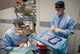 U.S. Air Force Col. (Dr.) Richard Townley, ophthalmologist at Joint Base Elmendorf- Richardson, Alaska, performs manual small incision cataract surgery on a patient while U.S. Air Force Maj. (Dr.) Zack Heath, ophthalmologist at Wilford Hall Ambulatory Surgical Center, Joint Base San Antonio- Lackland, Texas, stands by a mayo cart full of products and tools necessary for surgery. Both men are wearing gloves, an apron, mask and cap  to enhance operating room sterilization.