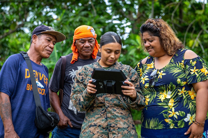 A Marine holds a mobile device as people in civilian clothing watch the screen.