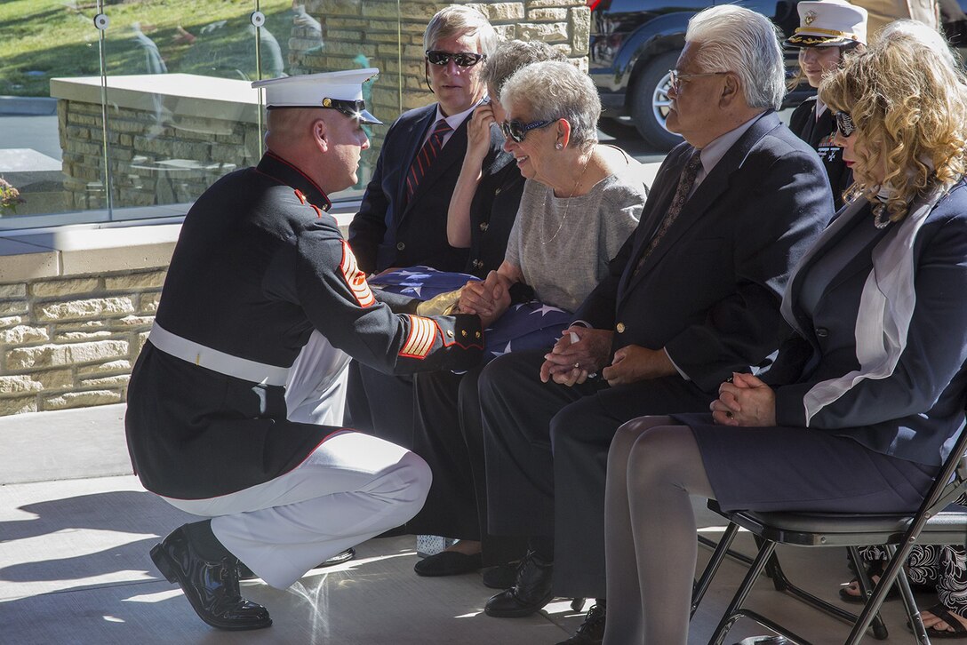 A uniformed service member kneels to talk to and hand a seated person a folded U.S. flag during a funeral service as other seated people watch.