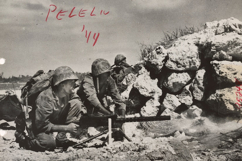 Service members crouch down to work a machine gun beside a pile of rocks.