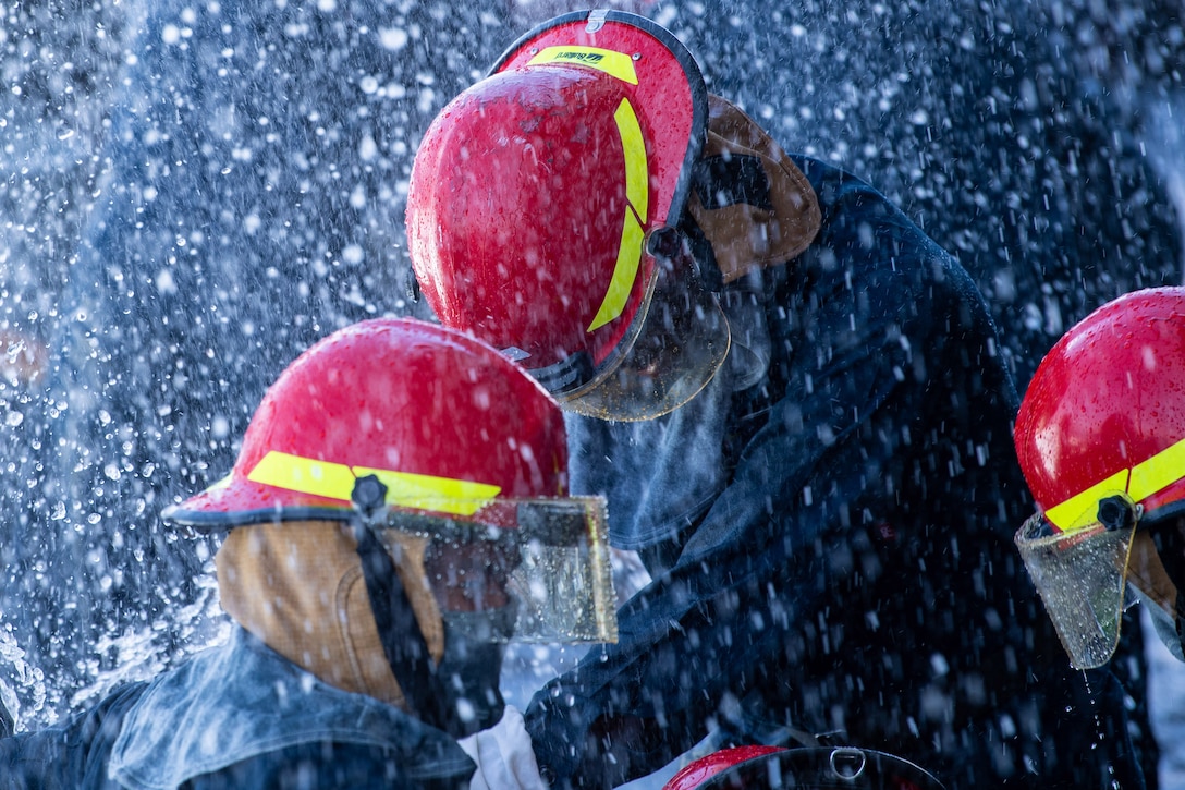 Water surrounds three sailors wearing fire gear including red helmets.