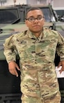 A young black man in U.S. Army camo military uniform leans back against the hood of a military vehicle. He has a close-cut hairstyle and black rectangular glasses with a slightly serious smile on his face, even as his body is relaxed against the vehicle. He's leaning on his elbows, propped on the hood.