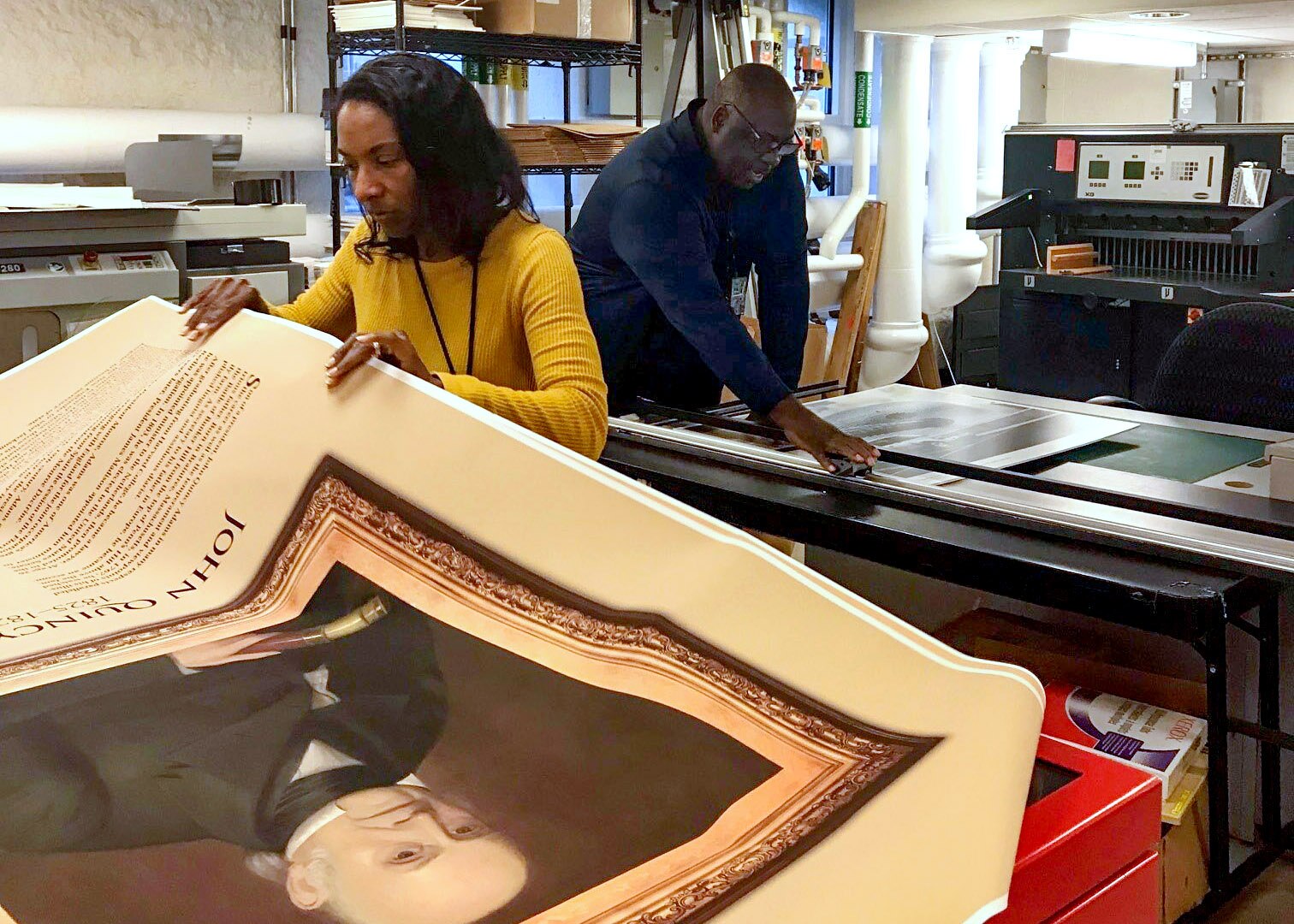 A woman looks at a large poster of a past president while the man behind her operates another print machine.