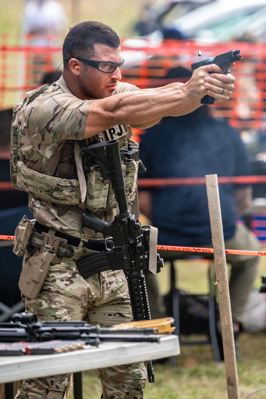 A service member aims a weapon while standing next to a table of guns.